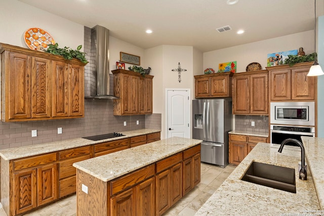 kitchen with a center island with sink, sink, light stone counters, appliances with stainless steel finishes, and wall chimney range hood