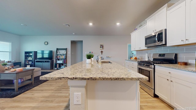 kitchen with stainless steel appliances, white cabinetry, light stone counters, a kitchen island with sink, and light hardwood / wood-style flooring