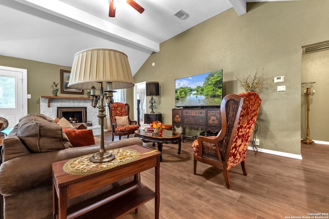 living room with ceiling fan, wood-type flooring, a stone fireplace, and vaulted ceiling with beams