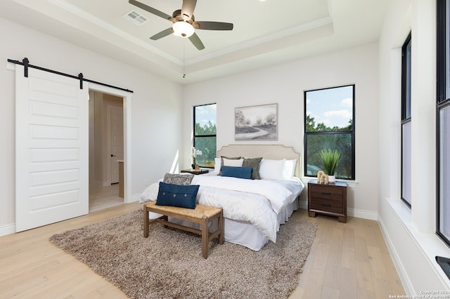 bedroom featuring light hardwood / wood-style floors, a barn door, ensuite bathroom, ceiling fan, and a tray ceiling