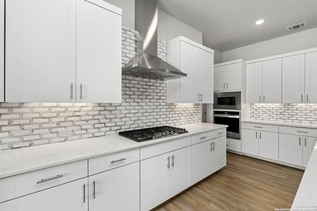 kitchen featuring stainless steel appliances, tasteful backsplash, wall chimney range hood, white cabinetry, and light wood-type flooring