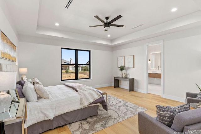 bedroom featuring a raised ceiling, ensuite bath, ceiling fan, and light wood-type flooring