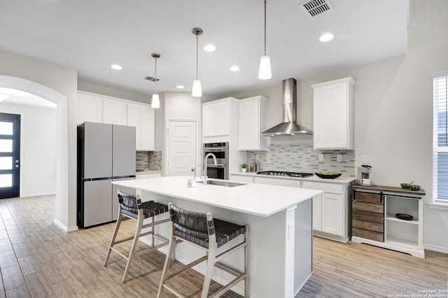 kitchen with appliances with stainless steel finishes, decorative light fixtures, wall chimney exhaust hood, and light hardwood / wood-style flooring