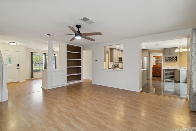 unfurnished living room featuring ornate columns, ceiling fan with notable chandelier, wine cooler, and light hardwood / wood-style flooring