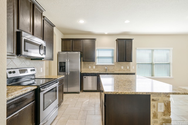 kitchen featuring light stone countertops, appliances with stainless steel finishes, tasteful backsplash, a kitchen island, and dark brown cabinetry