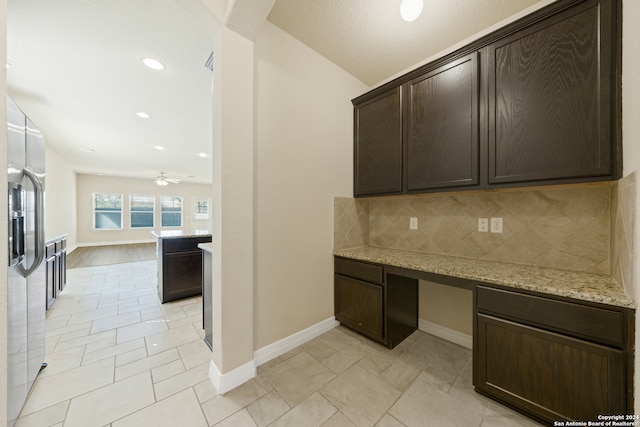 kitchen featuring decorative backsplash, ceiling fan, built in desk, dark brown cabinets, and light stone counters