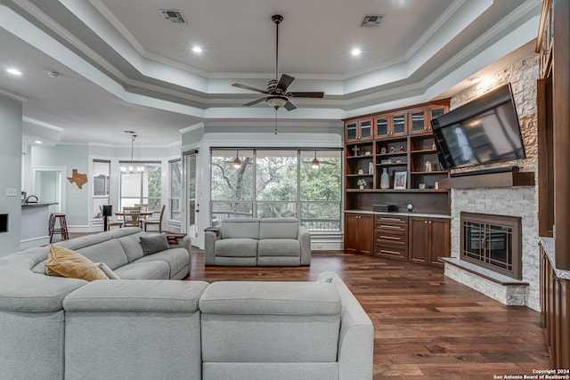 living room featuring a stone fireplace, ornamental molding, dark hardwood / wood-style floors, ceiling fan, and a tray ceiling