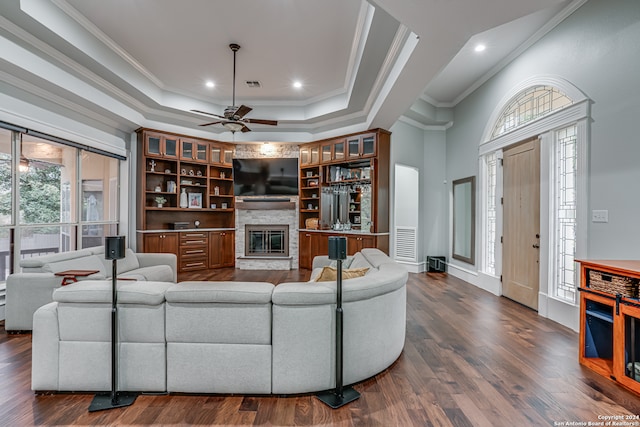 living room with ornamental molding, ceiling fan, dark hardwood / wood-style floors, a stone fireplace, and a raised ceiling