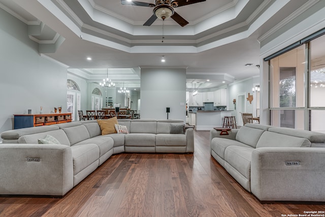 living room featuring dark wood-type flooring, a raised ceiling, ornamental molding, and ceiling fan with notable chandelier