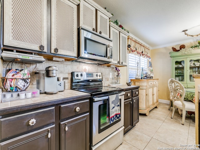 kitchen featuring stainless steel appliances, tile countertops, dark brown cabinetry, ornamental molding, and decorative backsplash