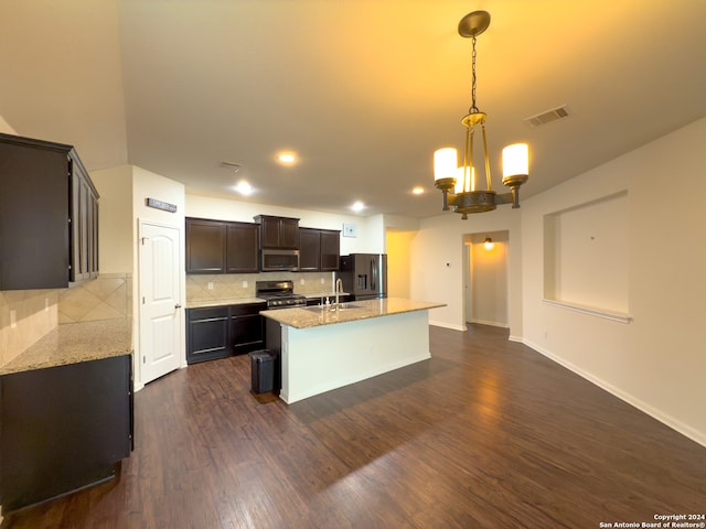 kitchen featuring dark hardwood / wood-style flooring, a center island with sink, light stone counters, appliances with stainless steel finishes, and decorative light fixtures