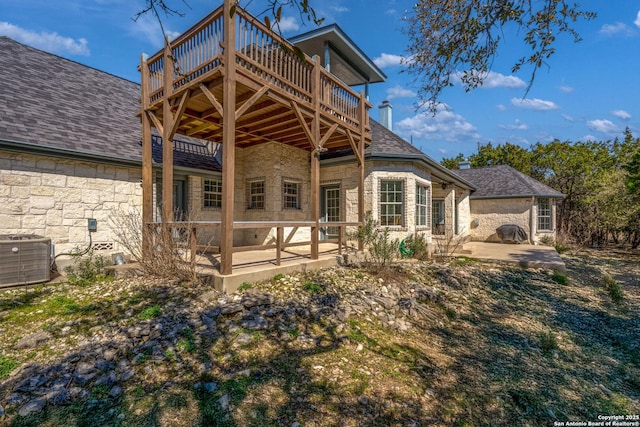 rear view of house with a deck, stone siding, roof with shingles, central AC unit, and a patio area