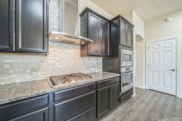 kitchen with wall chimney exhaust hood, light hardwood / wood-style flooring, stone counters, backsplash, and appliances with stainless steel finishes