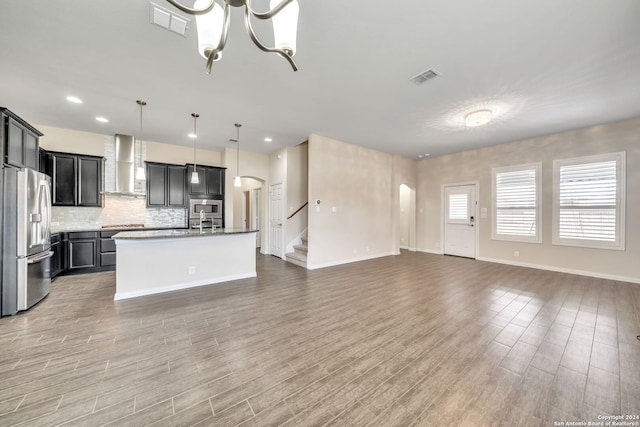 kitchen featuring stainless steel fridge with ice dispenser, hanging light fixtures, an island with sink, wall chimney exhaust hood, and light hardwood / wood-style flooring