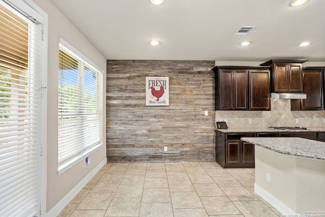 kitchen with decorative backsplash, dark brown cabinetry, light tile patterned floors, wooden walls, and light stone countertops