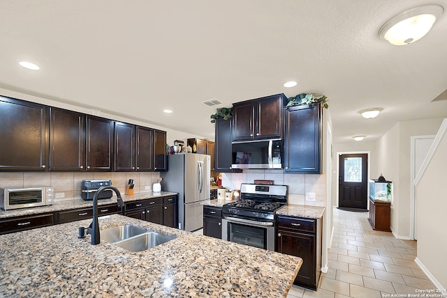 kitchen with sink, tasteful backsplash, light stone countertops, dark brown cabinets, and appliances with stainless steel finishes