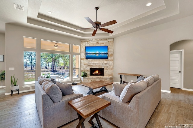 living room featuring light hardwood / wood-style flooring, a stone fireplace, ceiling fan, and a raised ceiling