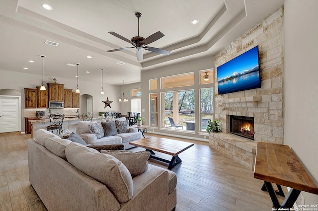 living room featuring a stone fireplace, ceiling fan, light hardwood / wood-style floors, and a raised ceiling