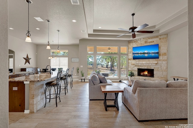 living room with a stone fireplace, light hardwood / wood-style floors, a textured ceiling, ceiling fan, and a tray ceiling