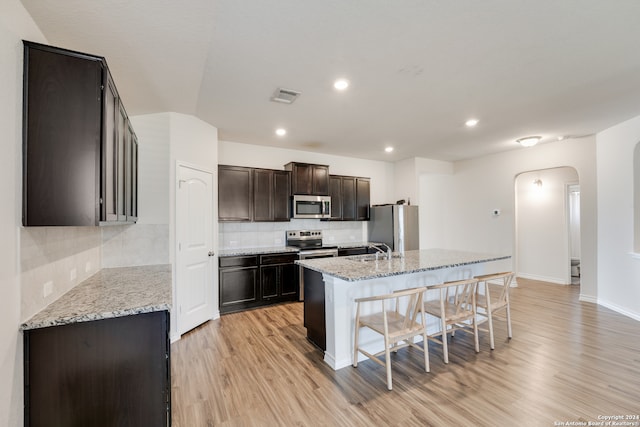 kitchen featuring light hardwood / wood-style floors, a center island with sink, light stone counters, appliances with stainless steel finishes, and tasteful backsplash