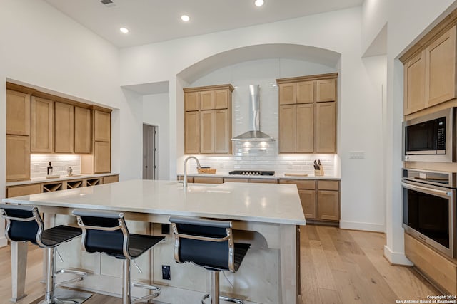 kitchen featuring stainless steel appliances, light wood-type flooring, sink, wall chimney exhaust hood, and a kitchen island with sink