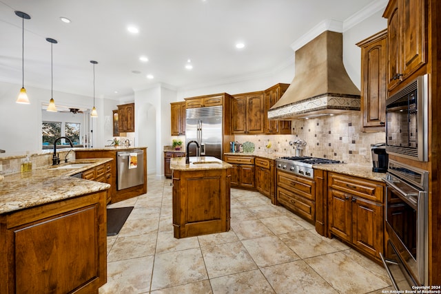 kitchen with built in appliances, hanging light fixtures, a kitchen island with sink, crown molding, and wall chimney range hood