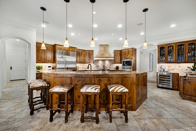 kitchen featuring beverage cooler, built in appliances, wall chimney exhaust hood, a large island with sink, and decorative light fixtures