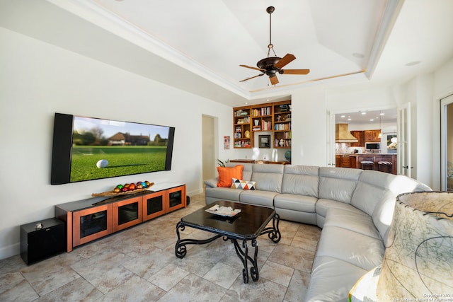 living room with ornamental molding, built in shelves, ceiling fan, and a raised ceiling