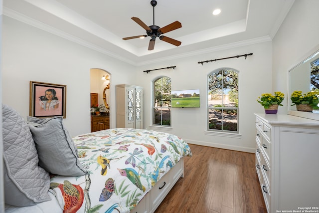 bedroom with crown molding, connected bathroom, dark hardwood / wood-style flooring, a tray ceiling, and ceiling fan