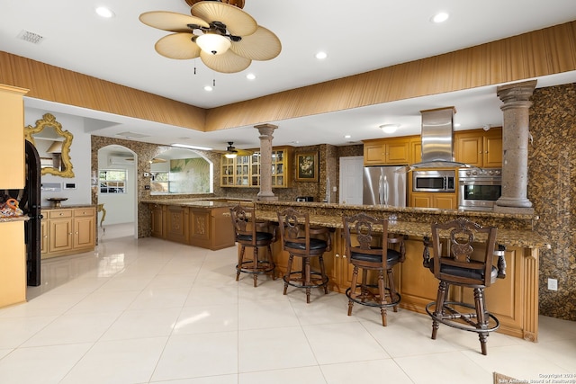 kitchen with ornate columns, stainless steel appliances, island range hood, kitchen peninsula, and a breakfast bar