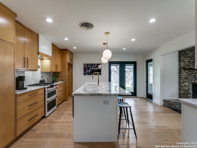 kitchen featuring decorative backsplash, light wood-type flooring, a kitchen island with sink, pendant lighting, and range with two ovens