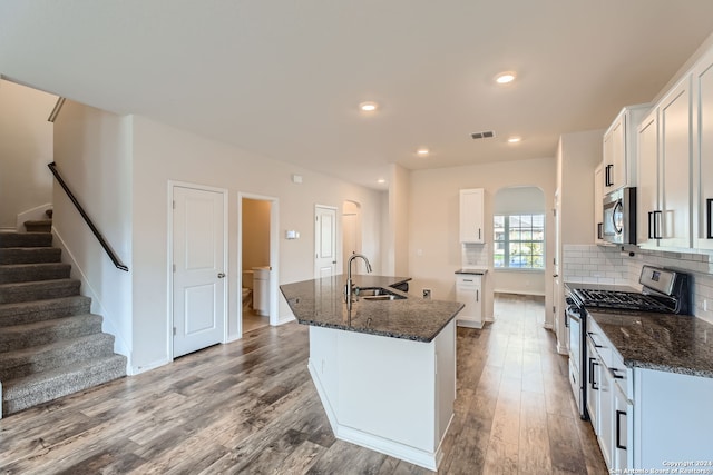 kitchen featuring white cabinetry, stainless steel appliances, light hardwood / wood-style flooring, dark stone countertops, and an island with sink