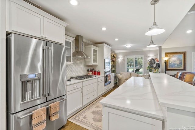 kitchen featuring white cabinets, appliances with stainless steel finishes, wall chimney exhaust hood, decorative light fixtures, and light stone counters