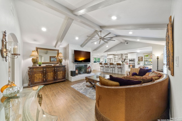 living room with a brick fireplace, lofted ceiling with beams, light hardwood / wood-style flooring, and ceiling fan