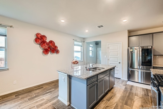 kitchen with stainless steel appliances, light stone counters, light hardwood / wood-style floors, and an island with sink