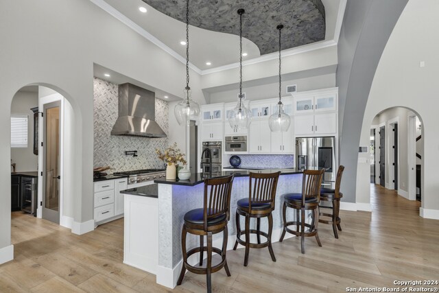 kitchen with wall chimney range hood, a center island with sink, a high ceiling, light hardwood / wood-style floors, and white cabinetry