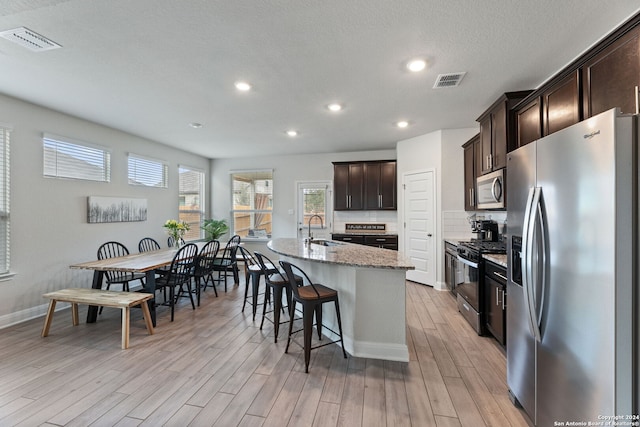 kitchen with light stone countertops, stainless steel appliances, sink, light hardwood / wood-style floors, and an island with sink