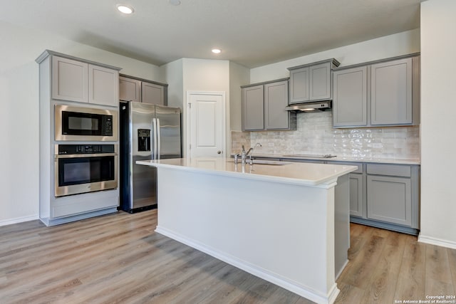 kitchen with appliances with stainless steel finishes, tasteful backsplash, sink, a center island with sink, and light hardwood / wood-style flooring