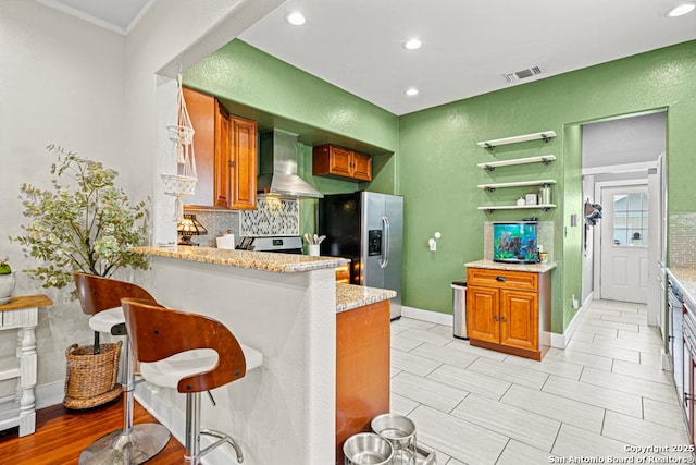 kitchen with a kitchen breakfast bar, wall chimney range hood, stainless steel refrigerator with ice dispenser, light stone counters, and kitchen peninsula