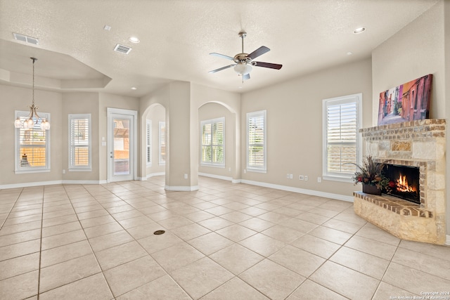 unfurnished living room featuring a stone fireplace, ceiling fan with notable chandelier, light tile patterned floors, and a textured ceiling