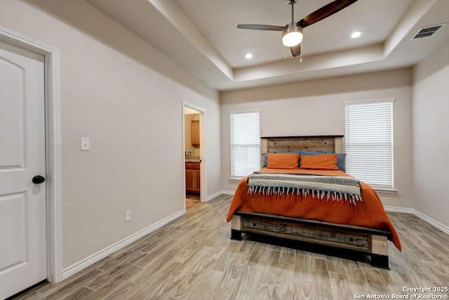 bedroom featuring ceiling fan, light wood-type flooring, ensuite bathroom, and a tray ceiling