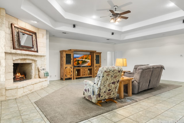 living room with ceiling fan, a stone fireplace, light tile patterned floors, and a tray ceiling