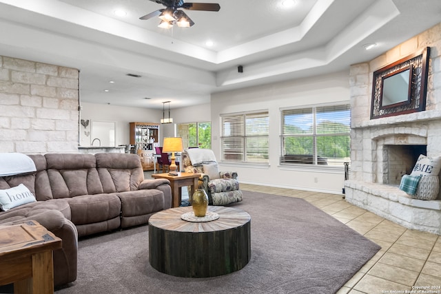 living room featuring light tile patterned floors, a raised ceiling, ceiling fan, and a stone fireplace