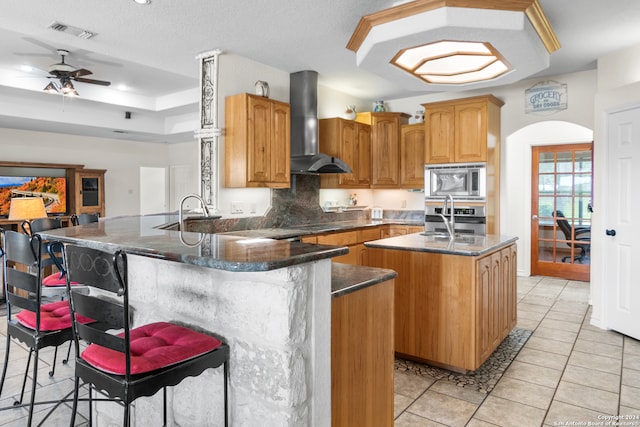 kitchen featuring kitchen peninsula, appliances with stainless steel finishes, wall chimney exhaust hood, a center island with sink, and a breakfast bar area