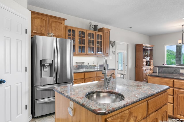 kitchen with sink, stainless steel fridge, dark stone counters, a textured ceiling, and a kitchen island with sink