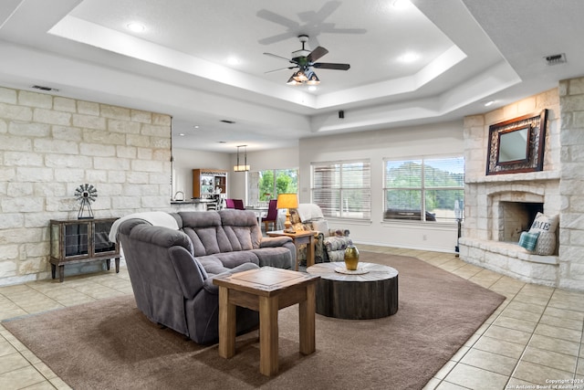 tiled living room featuring ceiling fan, a stone fireplace, and a tray ceiling