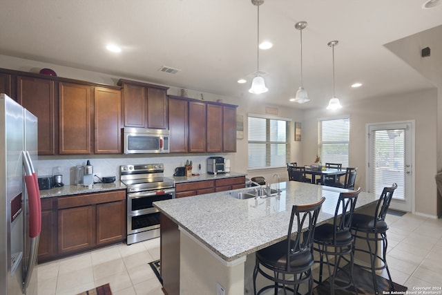 kitchen with sink, stainless steel appliances, light stone counters, pendant lighting, and a center island with sink