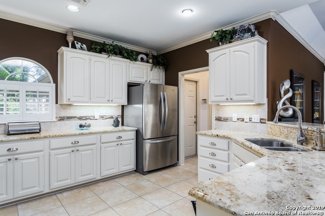 kitchen with stainless steel refrigerator, sink, light stone counters, white cabinets, and ornamental molding