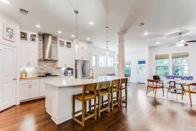 kitchen with dark wood-type flooring, hanging light fixtures, stainless steel appliances, wall chimney range hood, and a kitchen island with sink