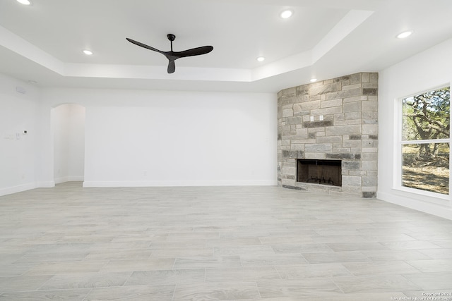 unfurnished living room with ceiling fan, a stone fireplace, and a tray ceiling